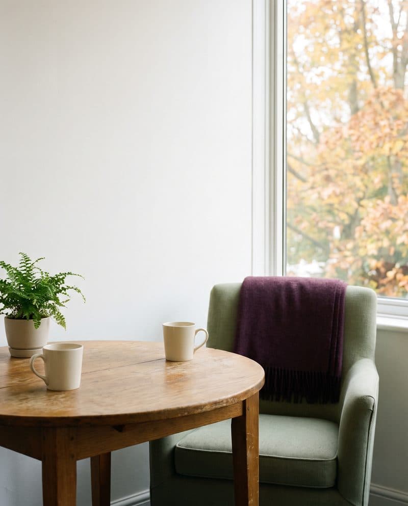 Two cream mugs on a round wooden table by a sunny window, with a sage armchair and a plum throw