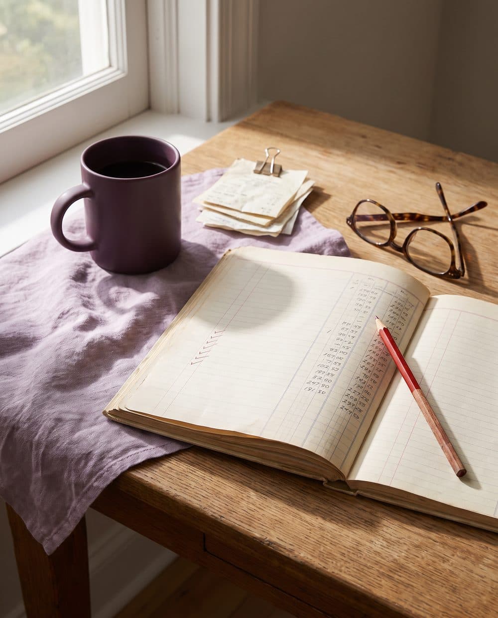 An open ledger with red pencil check marks, reading glasses and a plum coffee mug on an oak desk by a window